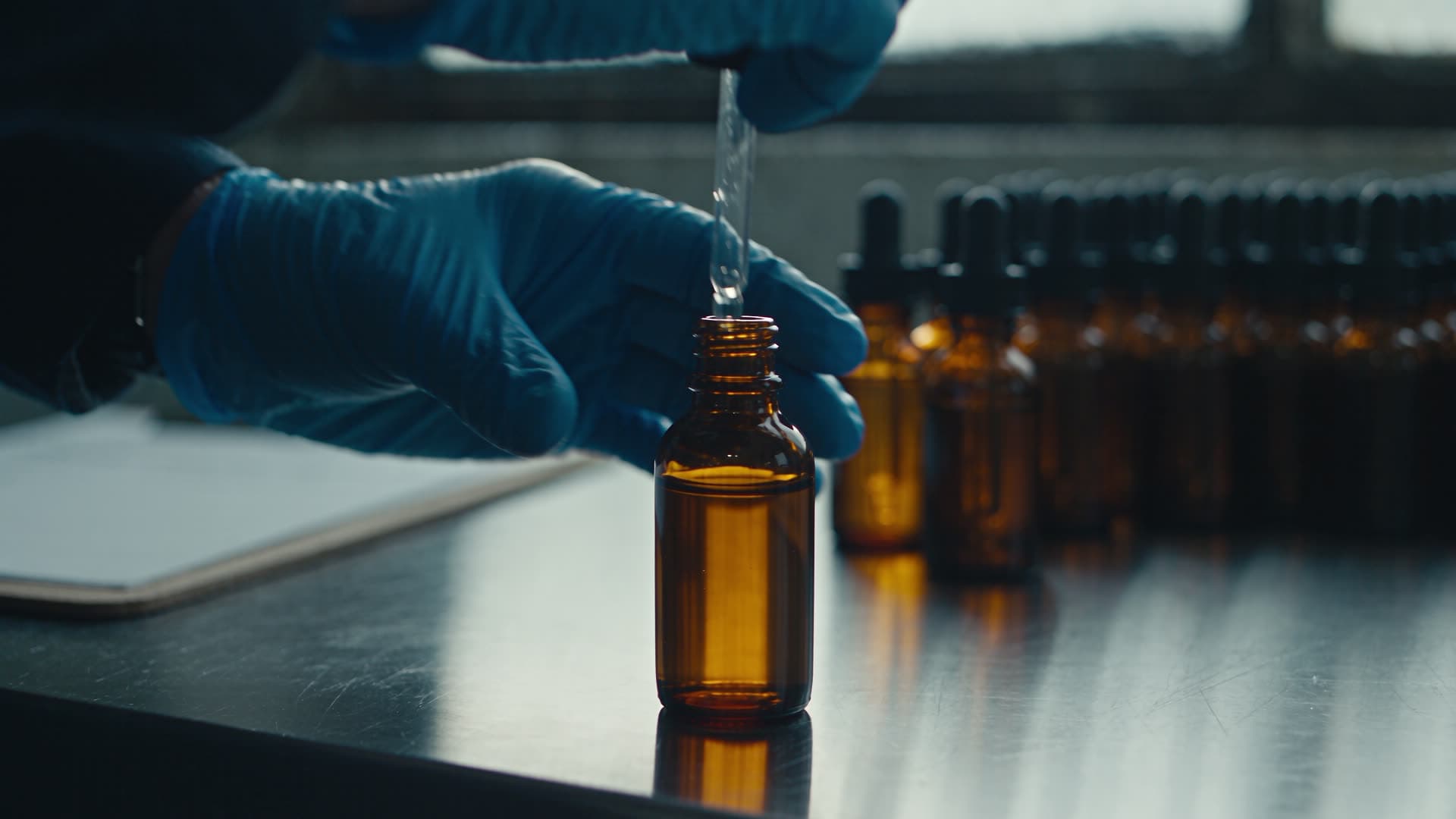 Gloved hands capping amber bottles on a steel bench in a small production room